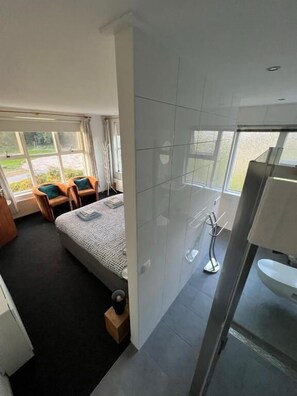 A modern hotel room with a large bed, a sitting area with a chair and table, a bathroom with a sink and mirror, and large windows offering a view of the outdoors. - B&B Zwiep (Lochem)