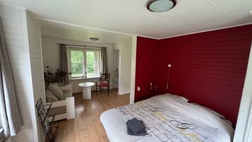 Et soveværelse med en stor seng, et lille bord og en stol. Et vindue med gardiner og udsigt til grøn bevoksning.