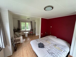 A bedroom with a large bed, a small table, and a chair. There is a window with curtains and a view of greenery outside. - B&B Zwiep (Lochem)