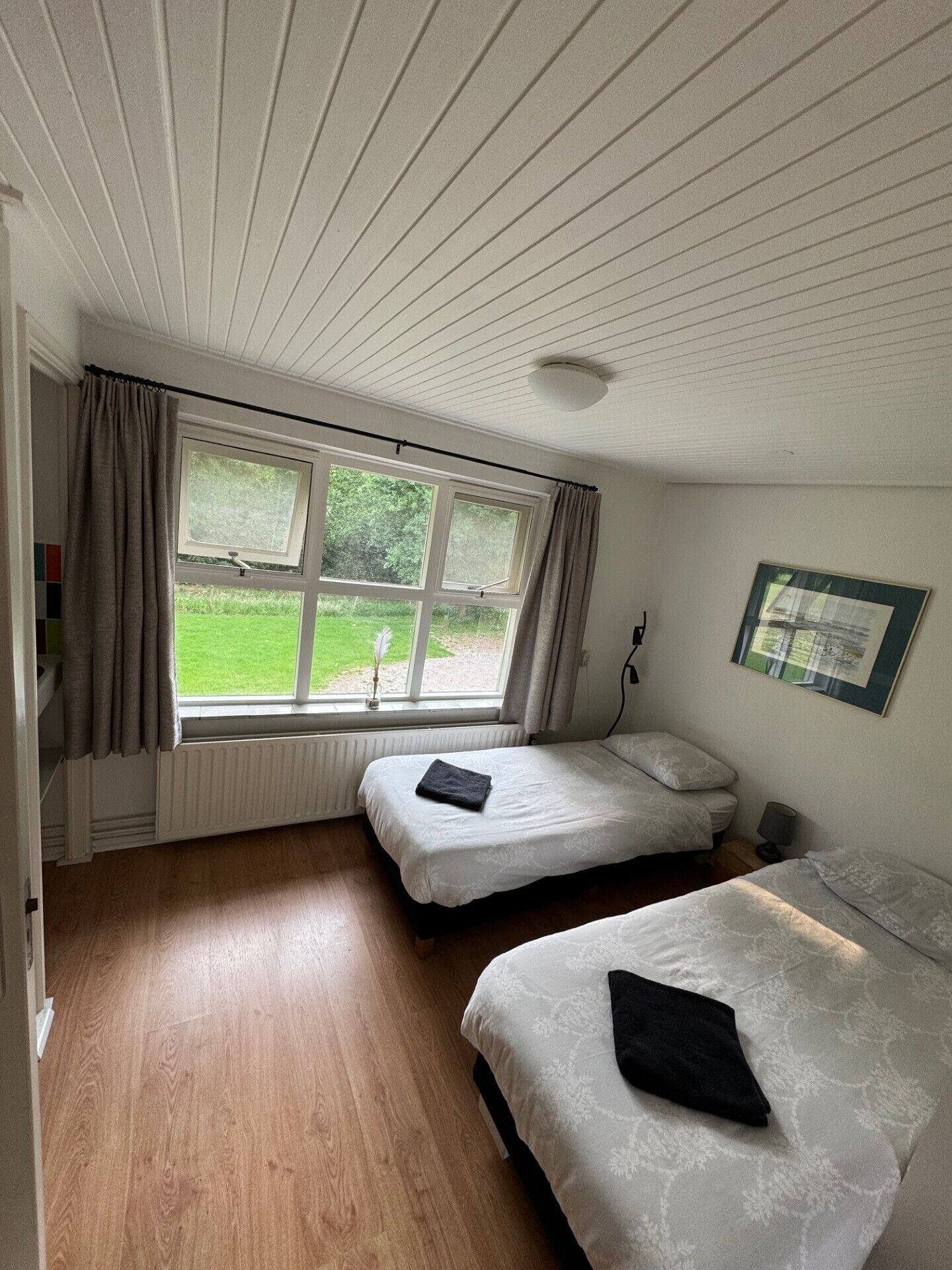 A room with two beds, a window with curtains, a radiator, and a framed picture on the wall.