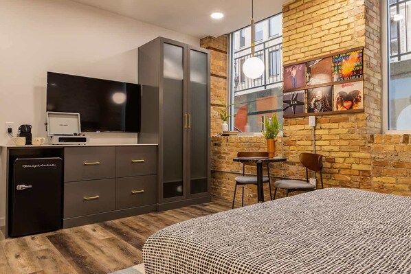 Una habitación de hotel moderna con una cama grande, un armario, un escritorio con computadora, un refrigerador pequeño, un televisor y una pared de ladrillo con obras de arte.