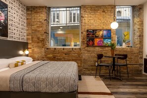 A modern hotel room with a brick wall, a large bed, a small table with chairs, and artwork on the wall. - 3 Private Boutique King Rooms - The Finnley Hotel (Grand Rapids)