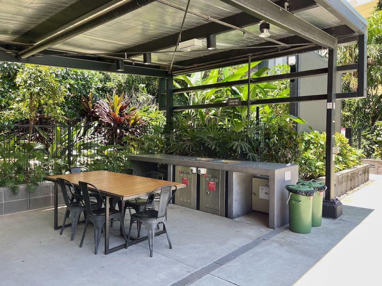 An outdoor seating area with a wooden table and chairs, a metal counter, and trash bins.
