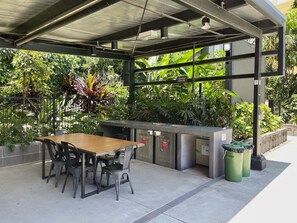 Un espacio al aire libre con mesas y sillas de madera, un mostrador de metal y contenedores de basura.