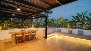 An outdoor patio with a wooden floor, a bar area with stools, a seating area with cushions, and a view of tropical plants and a palm tree. - 3-bedroom Aquamarine villa in awesome Bali with Private Pool (Bali)
