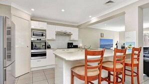 A modern kitchen with white cabinetry, a built-in oven, a microwave, and a large island with wooden chairs. - Hidden Horizon (Nords Wharf)