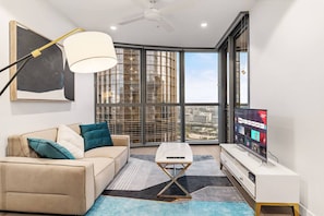 A modern living room with a sofa, a coffee table, a TV, and large windows offering a city view.