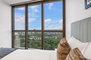 A bedroom with a large window, a bed with patterned pillows, and a view of a cityscape and greenery. - Budget Price for a Luxury Stay & Prime Facilities (Brisbane City)