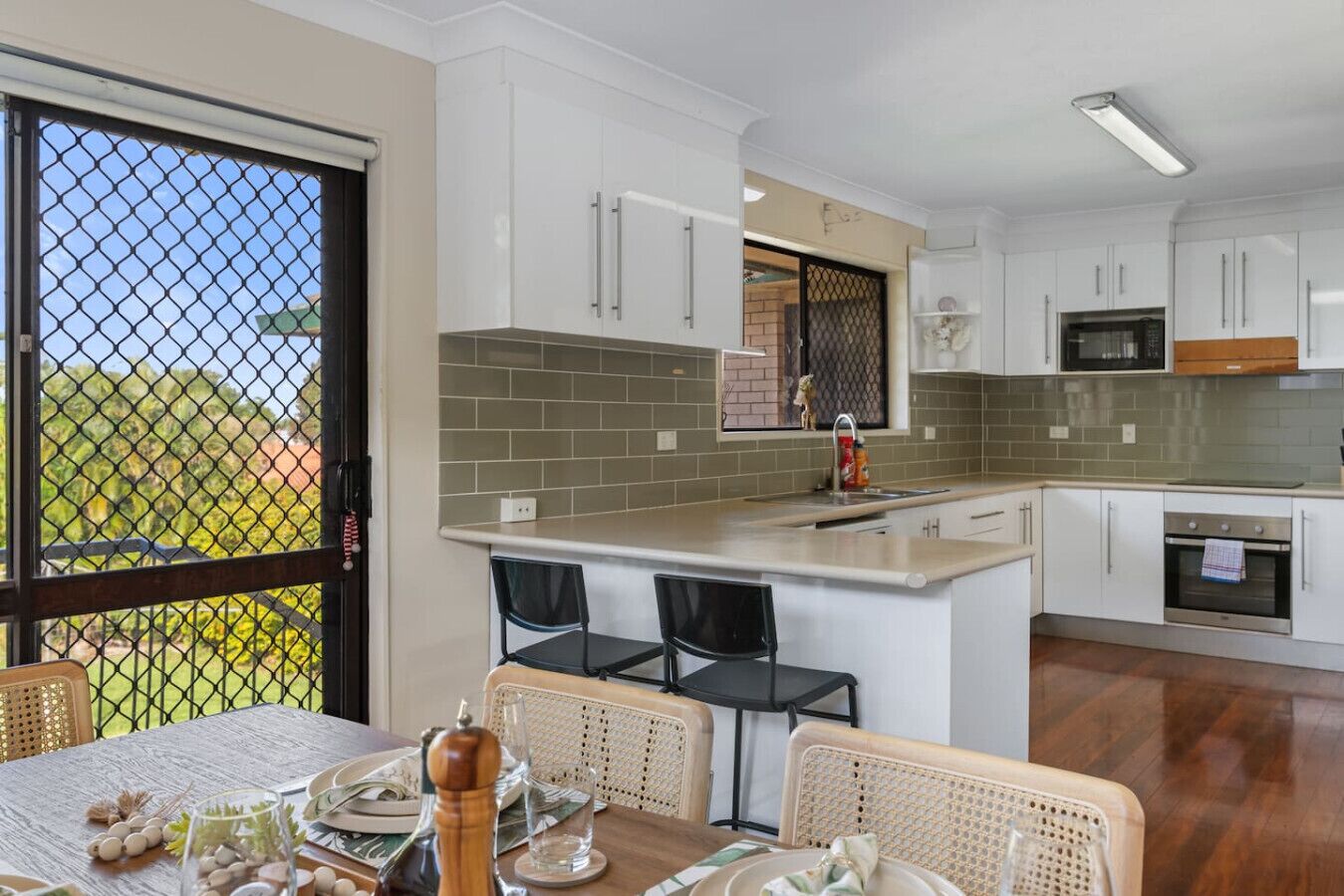 A modern kitchen with white cabinets, a grey tiled backsplash, and a built-in oven. There is a dining area with wicker chairs and a table set for two.
