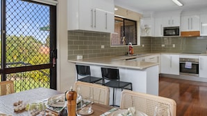 A modern kitchen with white cabinets, a grey tiled backsplash, and a built-in oven. There is a dining area with wicker chairs and a table set for two. - Unbeatable Value | Group/Family | Roomy/Convenient (Robertson)