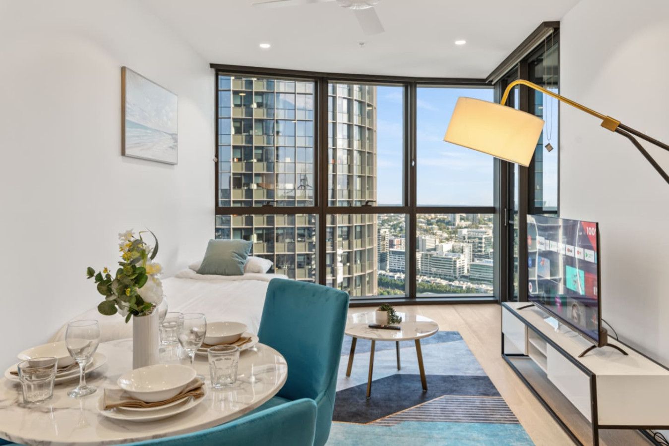 A modern hotel room with a large window, a bed, a dining area with a table set for two, and a TV on a stand.