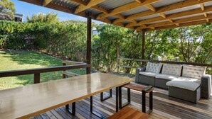 A covered outdoor patio with a wooden table and benches, a cushioned sofa, and a view of a lush garden. - 3Beds Value & Comfort Stay | Airport, CBD & Cafes (Bulimba)