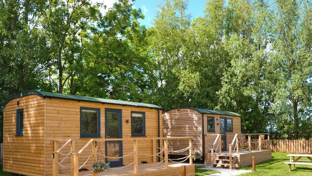 Una cabaña de madera con terraza, mesa de picnic y un área cercada con bancos.