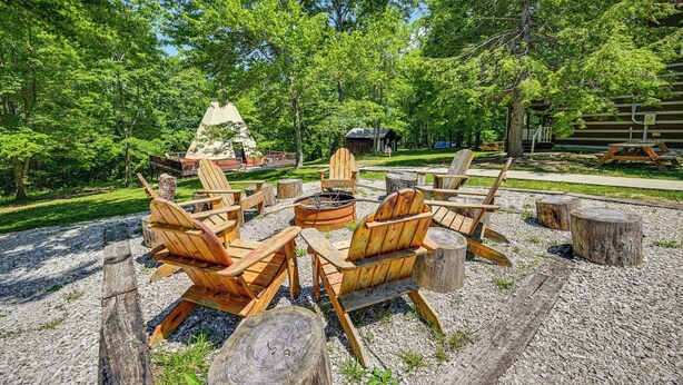 Ein rustikaler Außenbereich mit Holz-Adirondack-Stühlen und einem zentralen Feuerkorb, umgeben von Bäumen und einem Kiesweg.