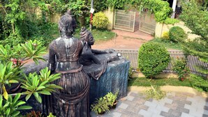 Una estatua de una figura sentada con un niño, rodeada de vegetación y un área pavimentada.