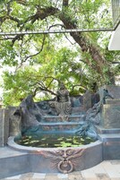 Una estatua de una figura sentada con intrincados grabados, un estanque con nenúfares y un árbol sobre ella.