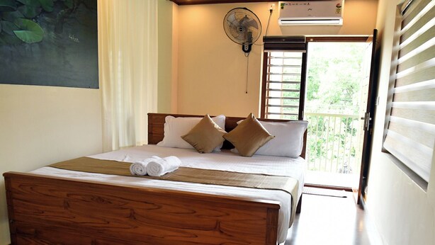 Una cama bien hecha con tablero de madera, ropa blanca y dos almohadas color beige. Hay un ventilador de pared sobre la cama, y se ve un equipo de aire acondicionado en la pared.