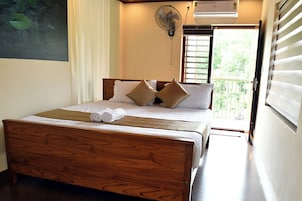 Una cama bien hecha con tablero de madera, ropa blanca y dos almohadas color beige. Hay un ventilador de pared sobre la cama, y se ve un equipo de aire acondicionado en la pared.