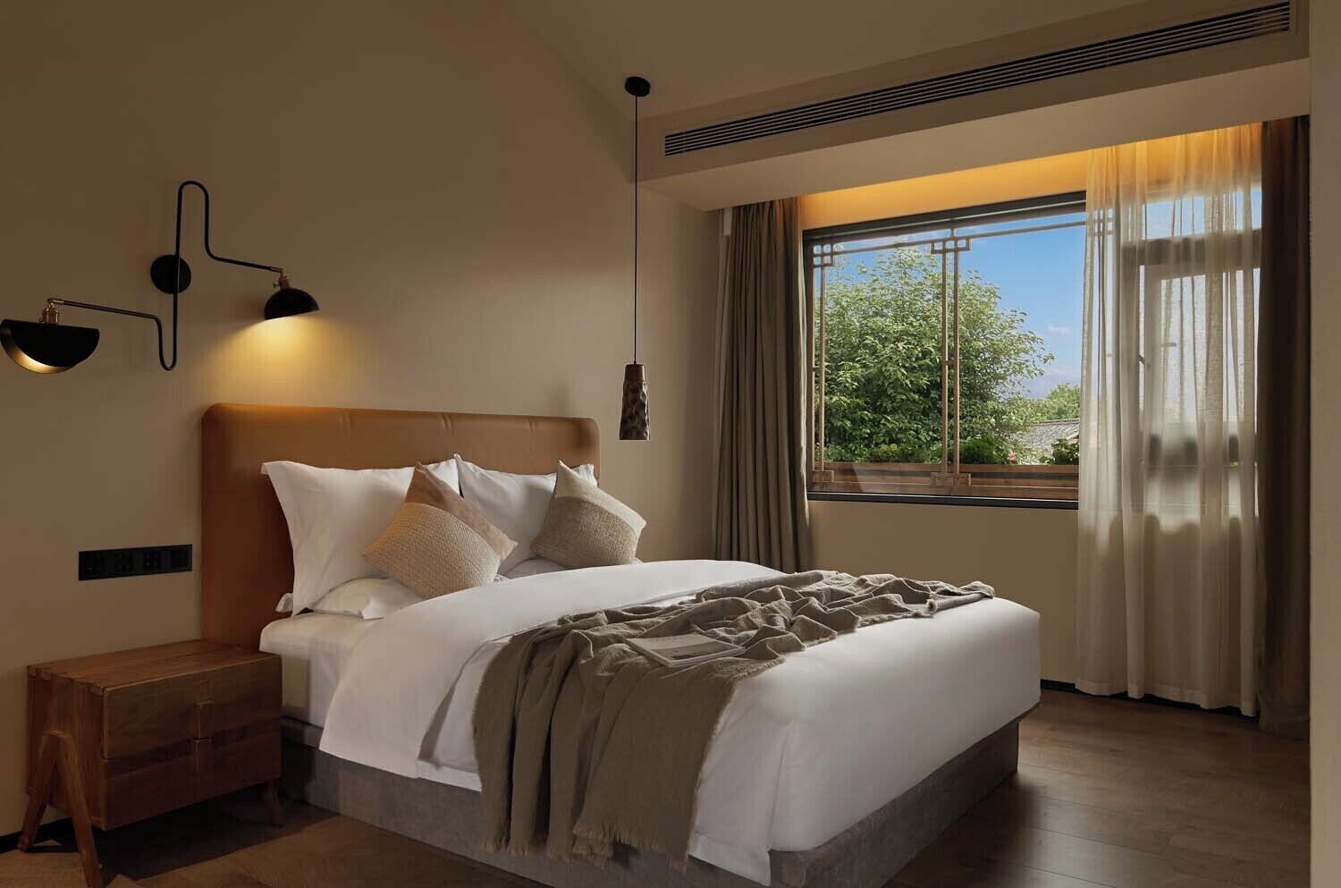 A modern hotel room with a large bed, a wooden nightstand, a wall-mounted lamp, and a window with a view of greenery.