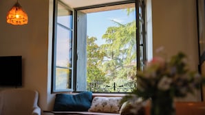 A room with a large window, a sofa with cushions, and a hanging light fixture. - Charming studio in lovely Les Vans, National Park Monts d'Ardèche, river nearby. (Les Vans)
