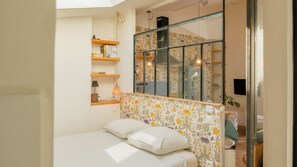 A bedroom with a bed, a shelf with books, a lamp, and a window with floral wallpaper.