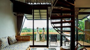 A room with a wooden sofa, a patterned rug, a small table with a book, and a view of two people outside through a glass door. - Oxy Resort Chiangrai (Mae Chan)