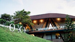 A building with a prominent 'OXY' sign on a grassy hill, featuring a modern architectural design with a brick facade and a sloped roof. - Oxy Resort Chiangrai (Mae Chan)