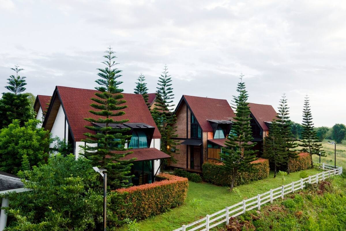 A row of modern houses with red roofs, surrounded by lush greenery and a white fence.