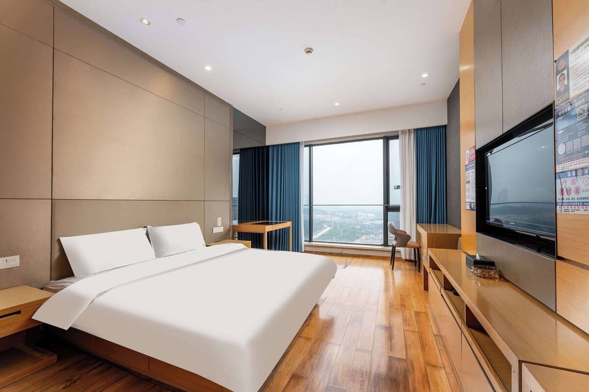 A modern hotel room with a large bed, wooden flooring, a TV, and a city view.