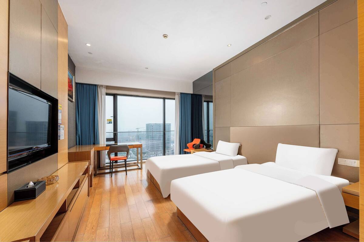 A hotel room with two beds, a desk, a chair, a television, and a large window with curtains.