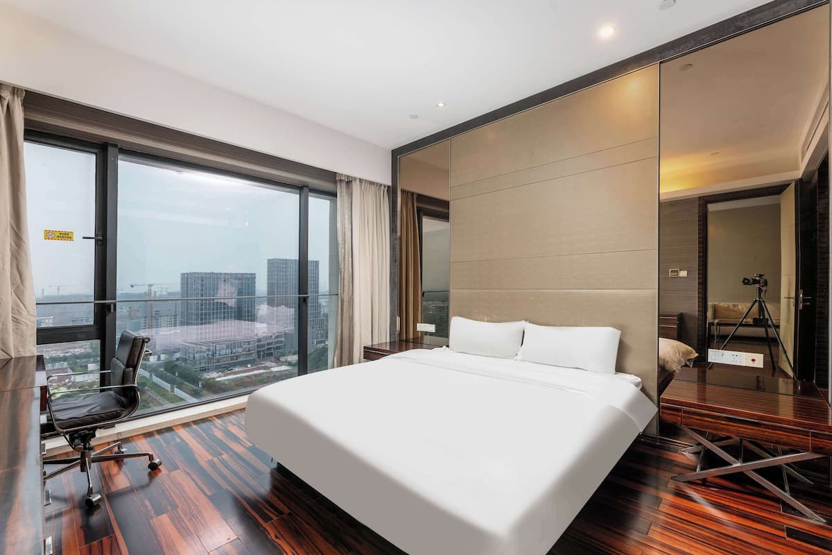 A modern hotel room with a large bed, a desk, and a chair. There is a large window with a view of the city.