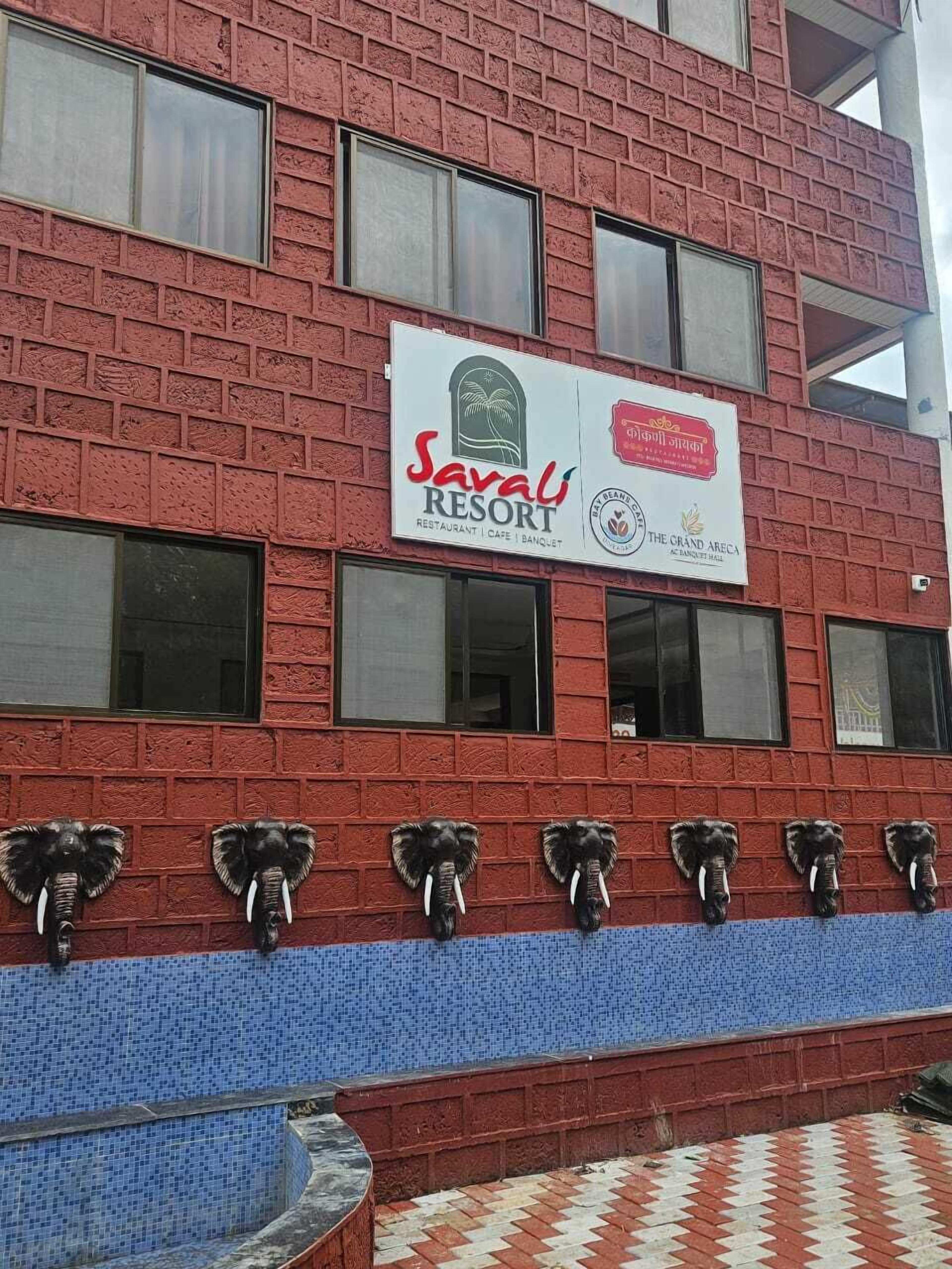 A building with a red brick facade, a sign for 'Savarli Resort' indicating it is a restaurant, cafe, and banquet hall, and decorative elephant head sculptures on the lower part of the facade.