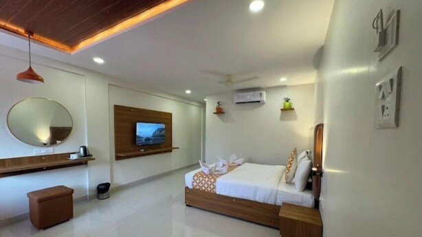 A modern hotel room with a wooden bed, a TV mounted on the wall, a round mirror, and a small wooden bench. - Savali Resort (Diveagar)