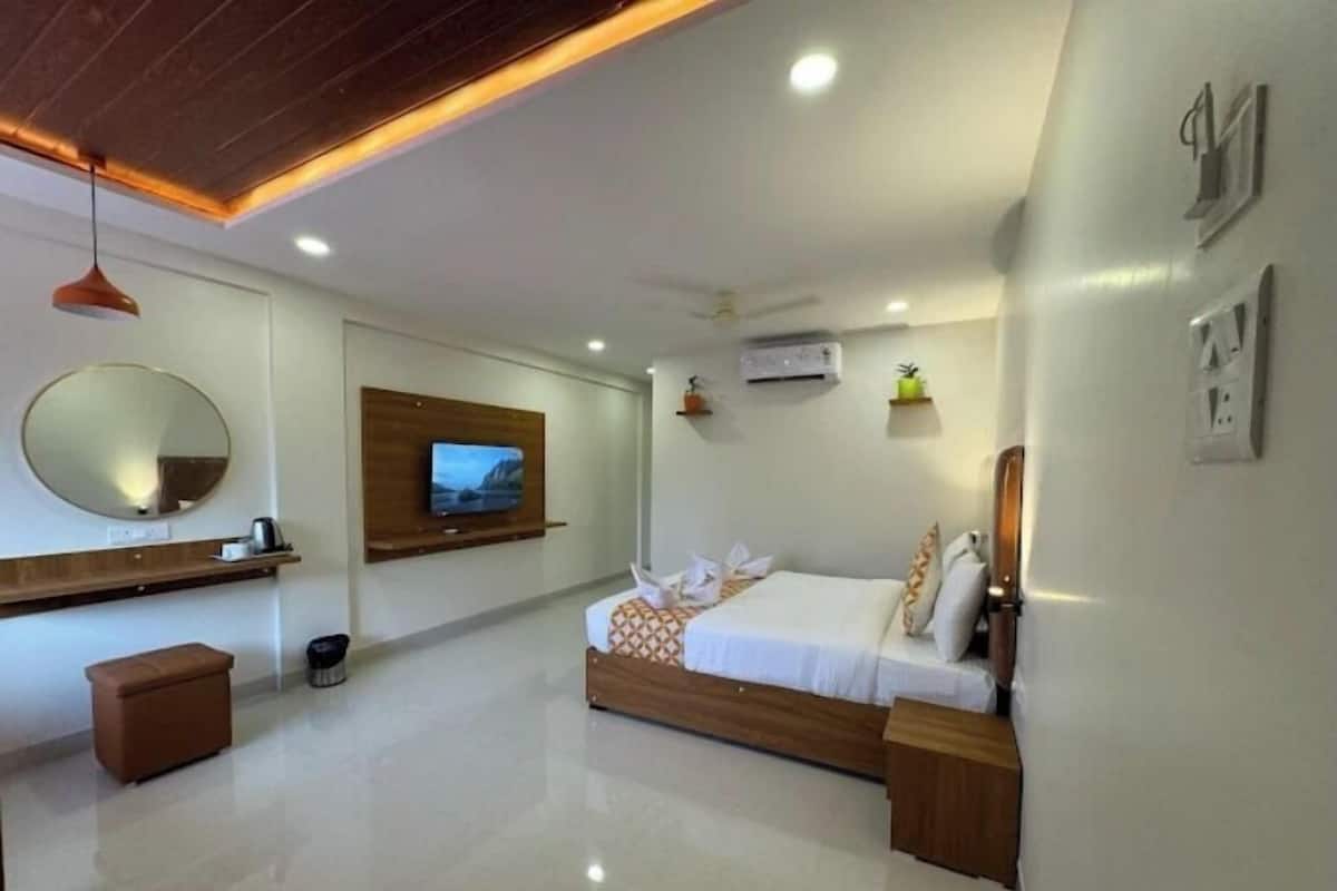 A modern hotel room with a wooden bed, a TV mounted on the wall, a round mirror, and a small wooden bench.
