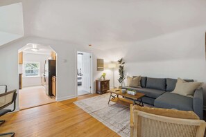 Uma sala de estar espaçosa com piso de madeira, um sofá cinza, uma mesa de centro de madeira e uma cadeira de rattan.
