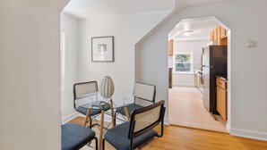 Un comedor con una mesa y sillas de vidrio, un cuadro en la pared y una cocina visible a través de un umbral abierto.
