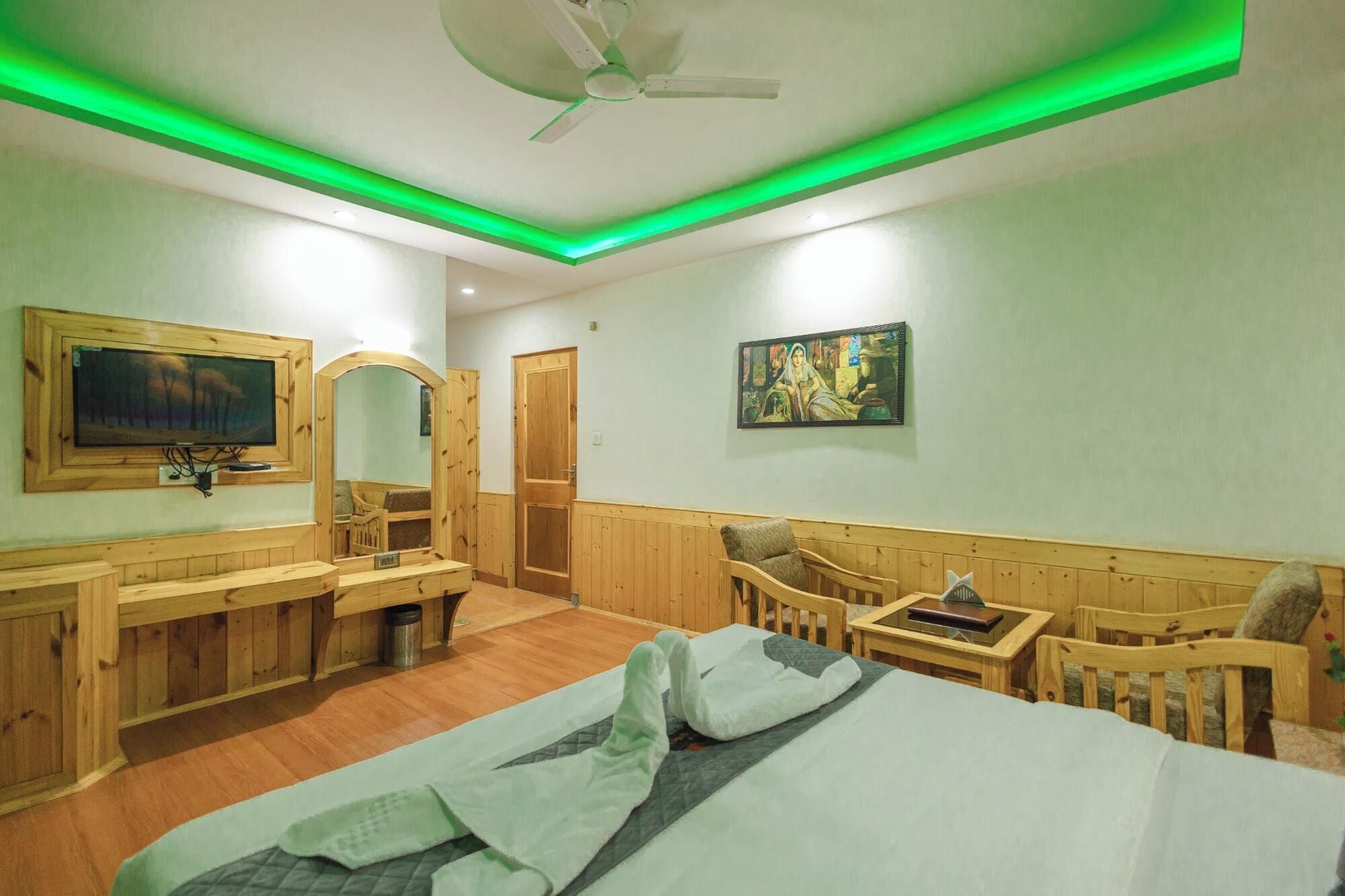 A hotel room with a bed, wooden furniture, a TV, a painting, and a ceiling fan.