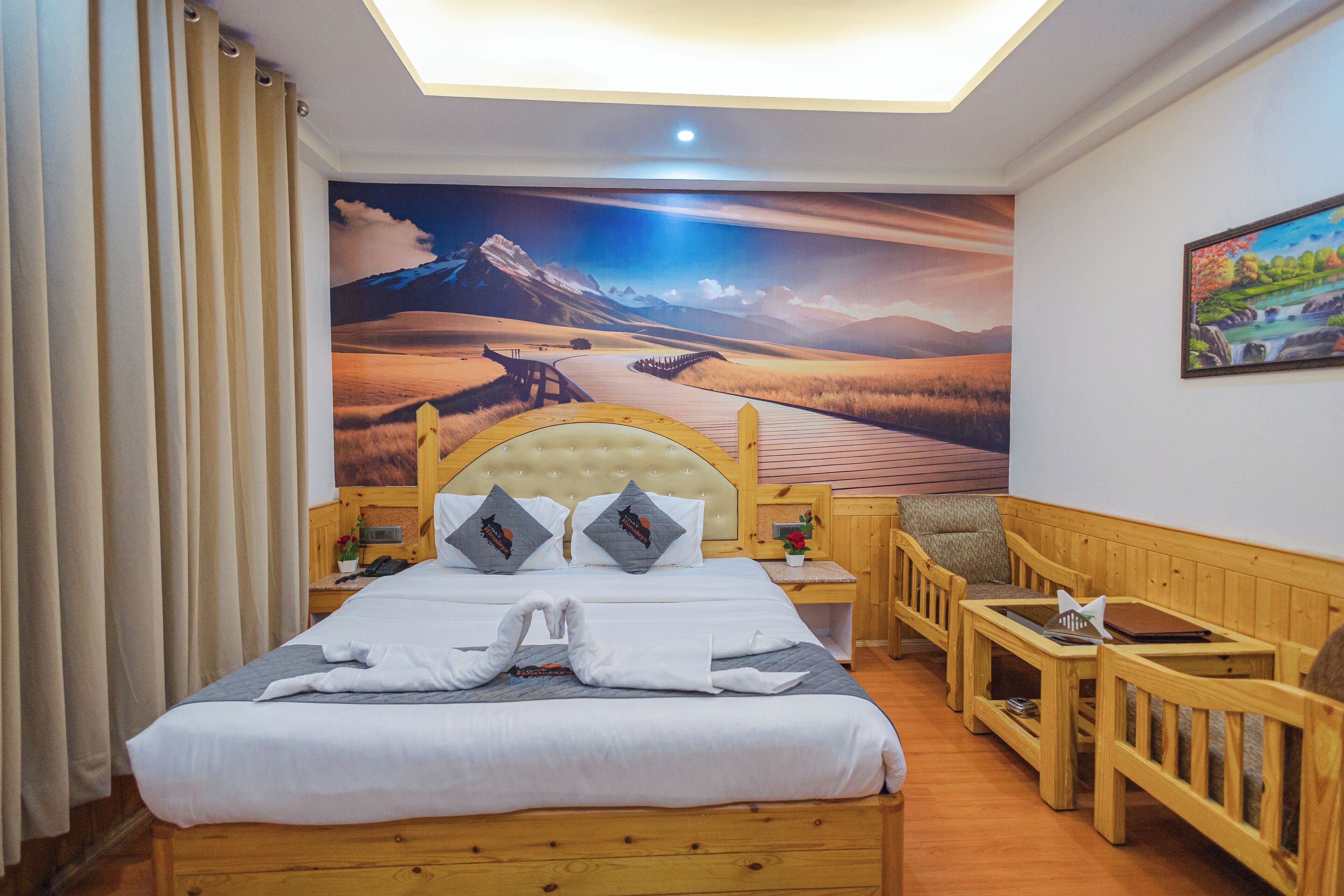 A hotel room with a large bed, wooden headboard, a mural of a landscape, a wooden bench, and a small table.