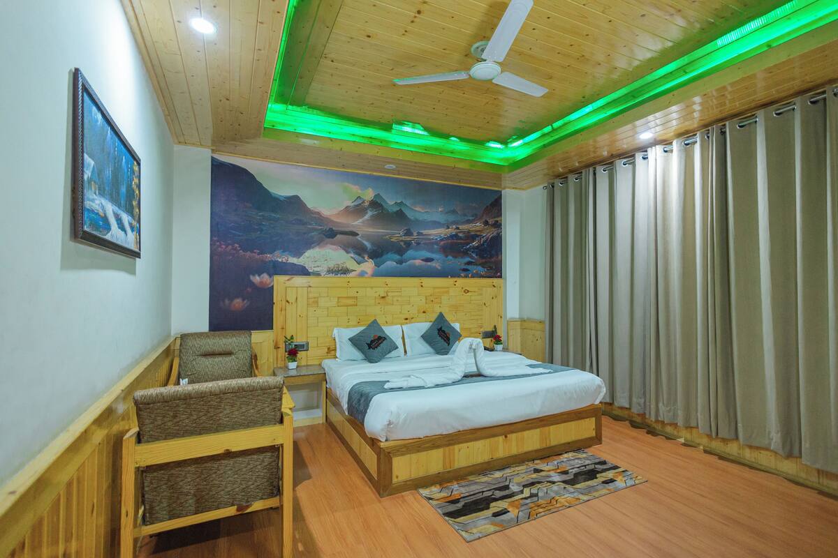 A bedroom with a wooden headboard, a bed with pillows, a nightstand, a chair, a large mural on the wall, and a window with curtains.