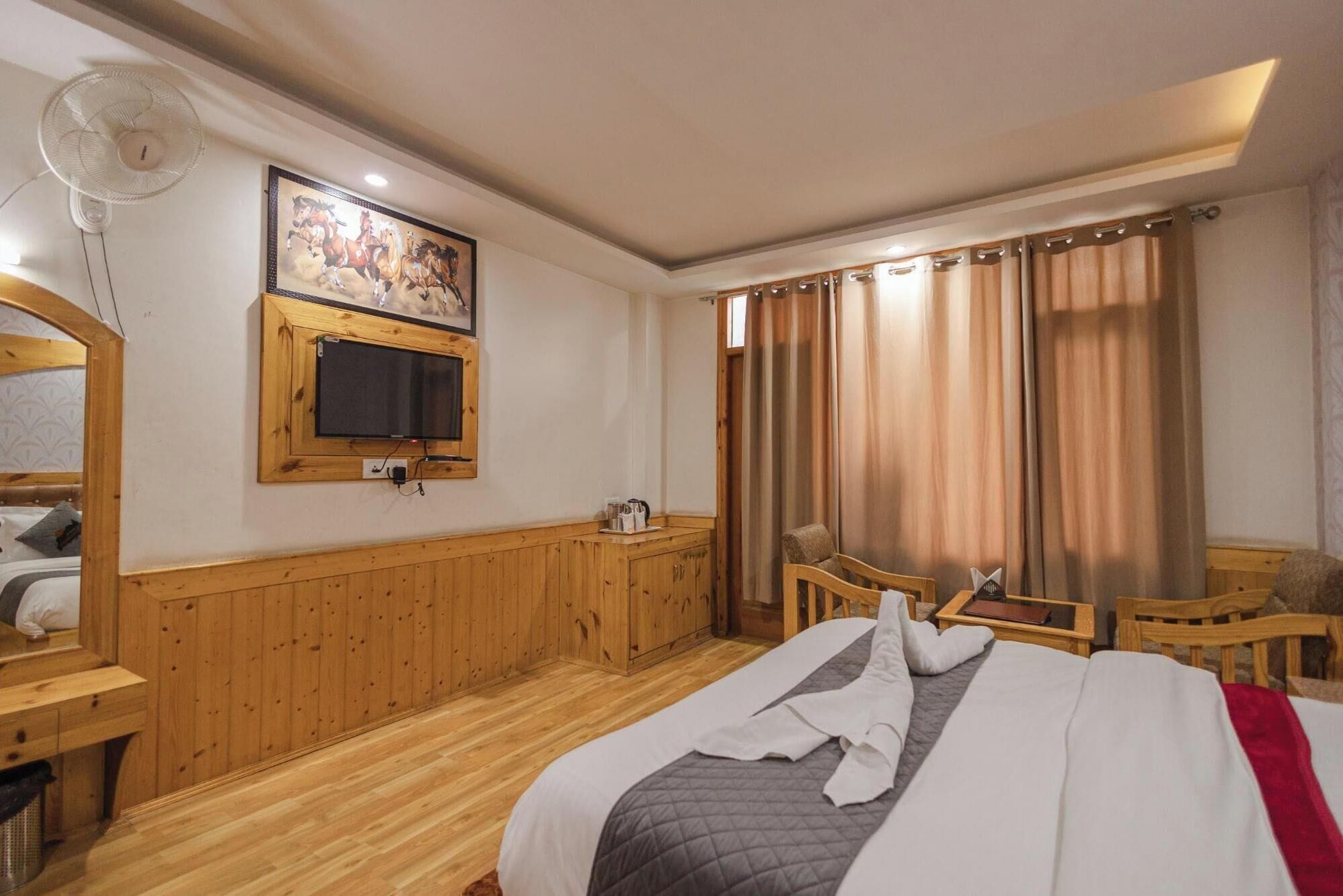 Ein Hotelzimmer mit einem Bett, einem an der Wand befestigten Fernseher, einem Holztisch, einem Stuhl und einem Fenster mit Vorhängen.