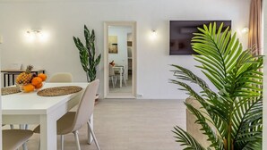 Uma sala de jantar moderna com uma mesa branca, cadeiras e uma fruteira. Há duas luminárias na parede e uma planta grande em primeiro plano.
