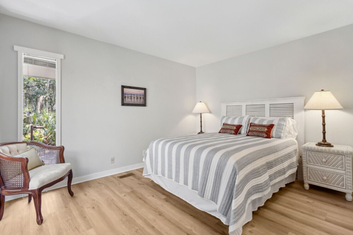 A bedroom with a large bed, two bedside tables with lamps, a chair, and a window with a view of greenery.