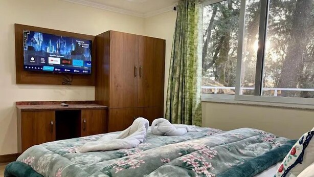 A bedroom with a bed, a TV mounted on the wall, and a window with curtains. - Jakhu heights (Shimla)
