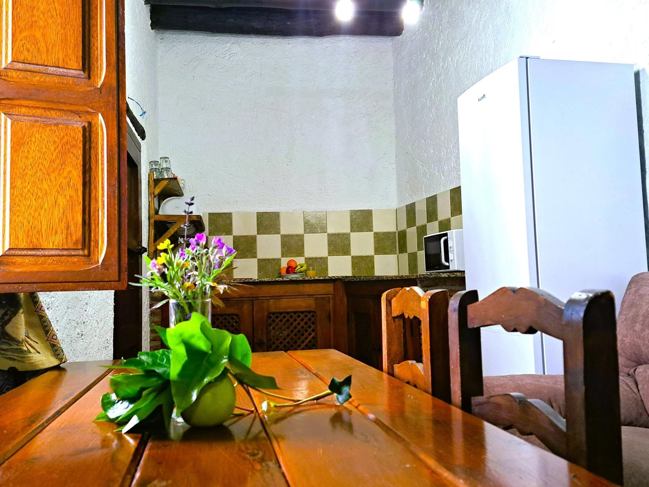 A wooden dining table with a vase of flowers, a microwave, and a refrigerator in a kitchen.