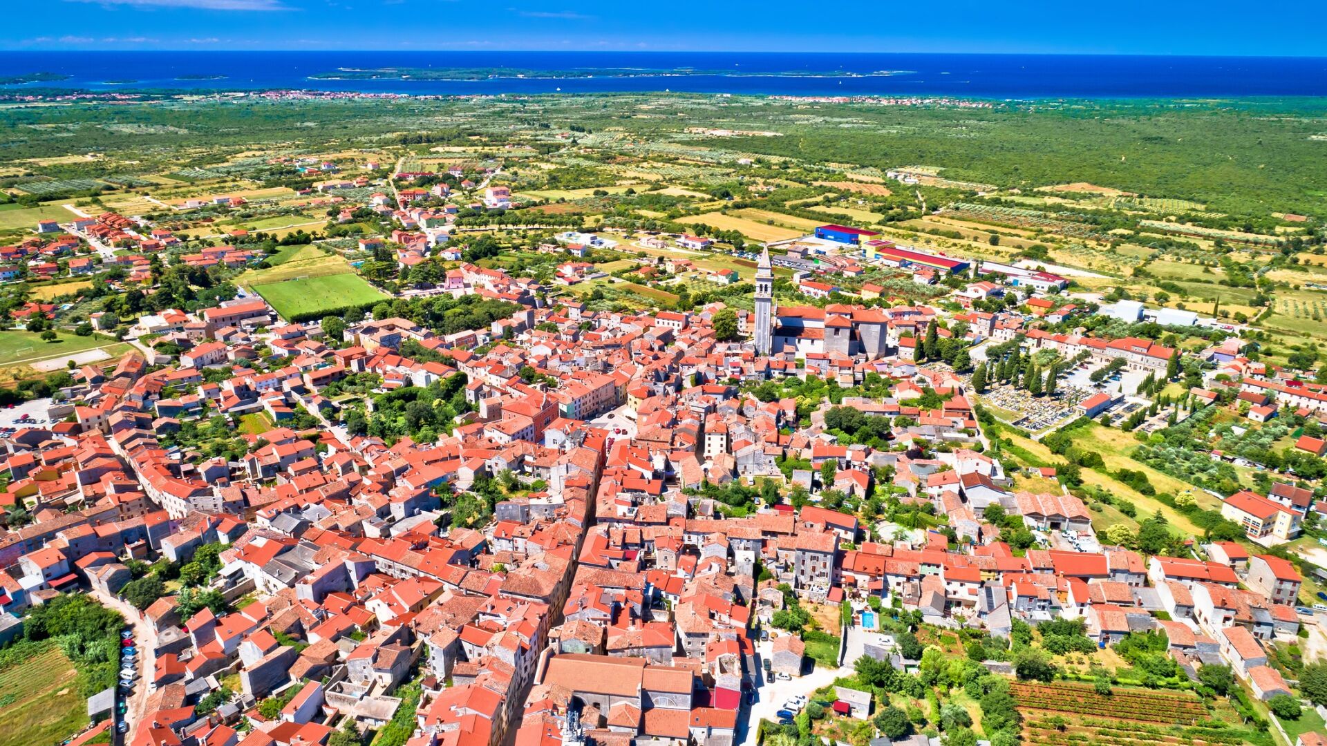 Une vue aérienne d’une ville avec des bâtiments aux toits rouges, une église imposante et des champs verdoyants environnants.