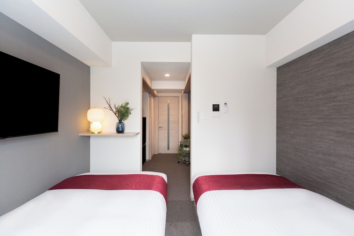 A hotel room with two beds, a TV mounted on the wall, a small plant on a shelf, and a hallway leading to the door.