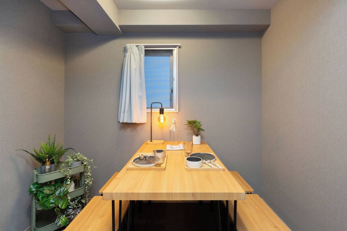 A compact room with a wooden table, a window with a curtain, a small plant, and a wall-mounted lamp.
