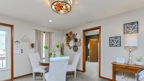 Comedor con una mesa redonda, dos sillas blancas, una mesita auxiliar de madera con una lámpara, y decoración en la pared que incluye platos y un letrero.