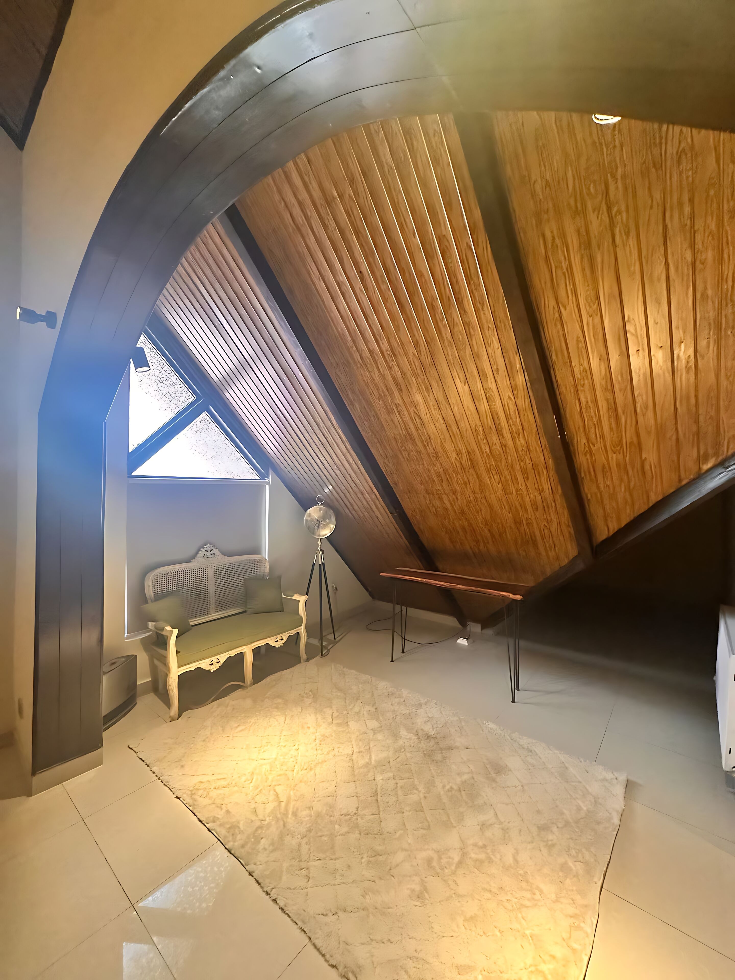Un dormitorio en el ático con techo inclinado, una banca de madera, una lámpara de pie y un ventanal grande.