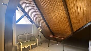 Un dormitorio en el ático con techo inclinado, una banca de madera, una lámpara de pie y un ventanal grande.